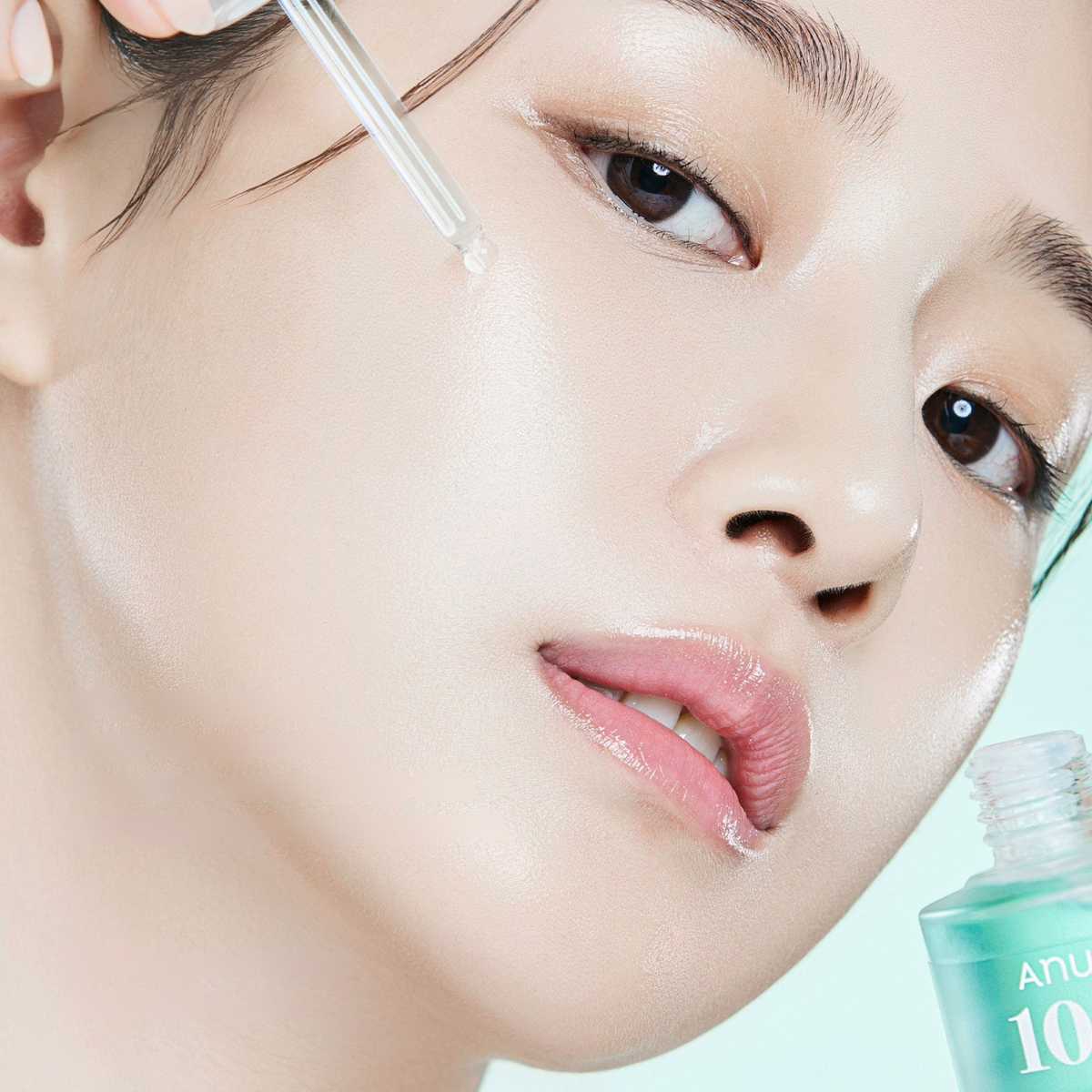Close-up of a woman's face with a glass dropper applying a clear skincare serum, with a turquoise bottle partially visible in the lower right corner.