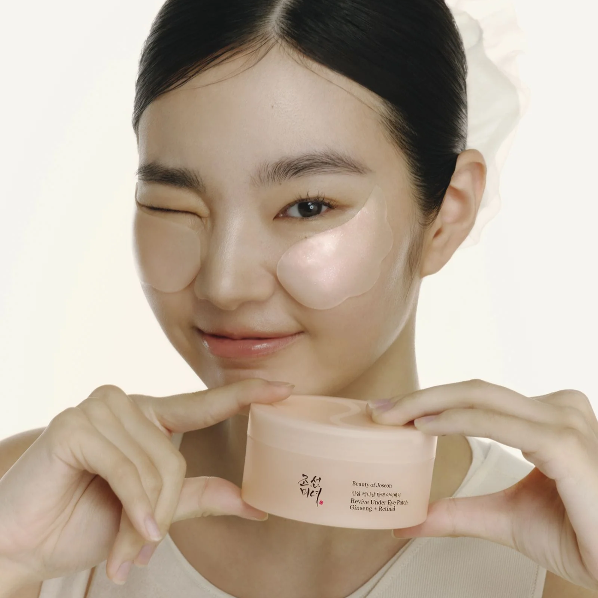 A woman with under-eye patches holds a small peach-colored cosmetic jar in front of her face against a clean white background.