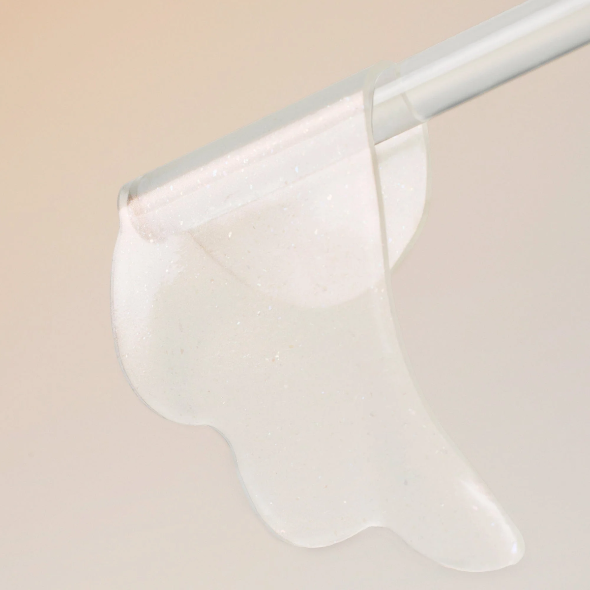 A close-up of a translucent, glittery clear cosmetic gel or adhesive being spread with a spatula on a soft beige background.