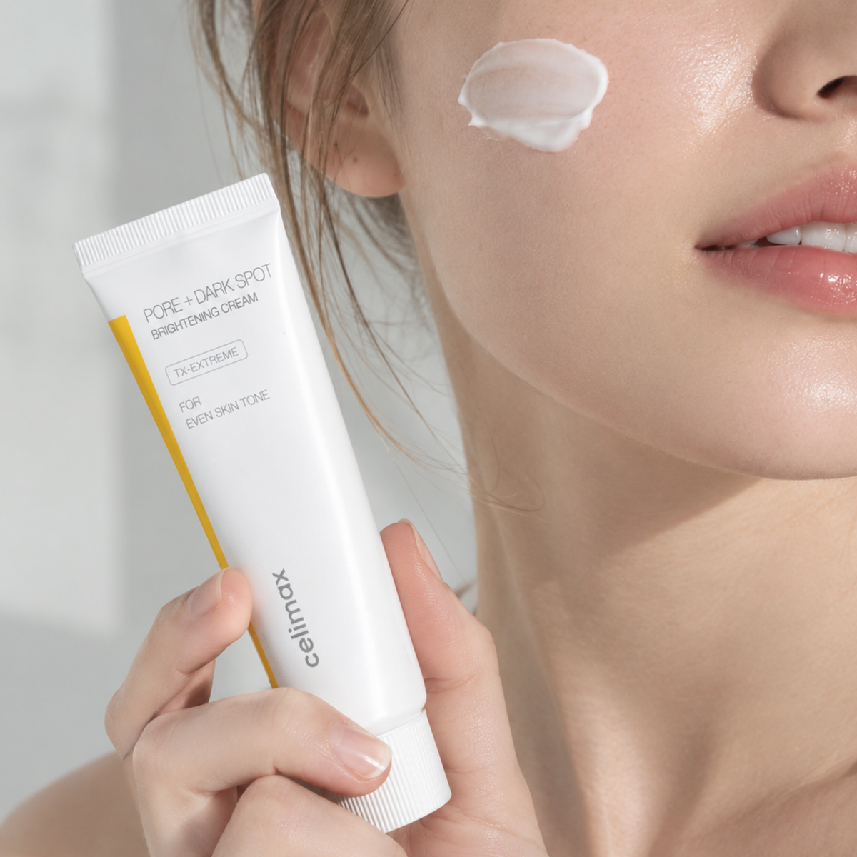 A close-up of a woman holding a white tube of Cellmax Pore + Dark Spot Brightening Cream, with a dollop of the white cream visible on her cheek against a soft, light background.