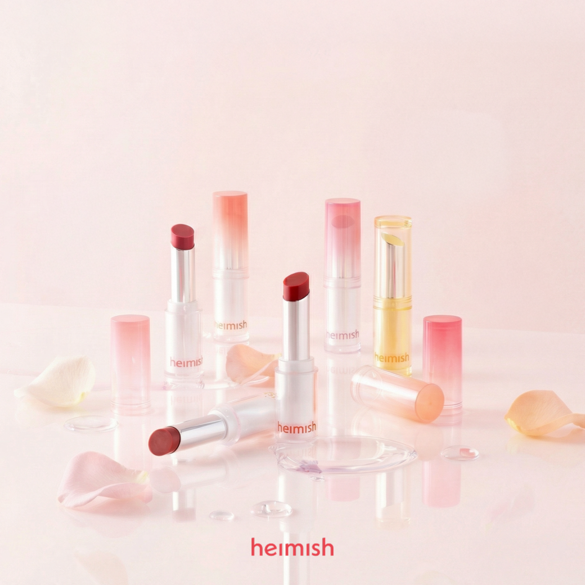 A group of Heimish lipsticks and lip balm tubes arranged on a glossy white surface with a soft pink background. Several tubes are open, showing deep berry-red and coral-pink balm colors, while others are closed in translucent pink and pale yellow cases.