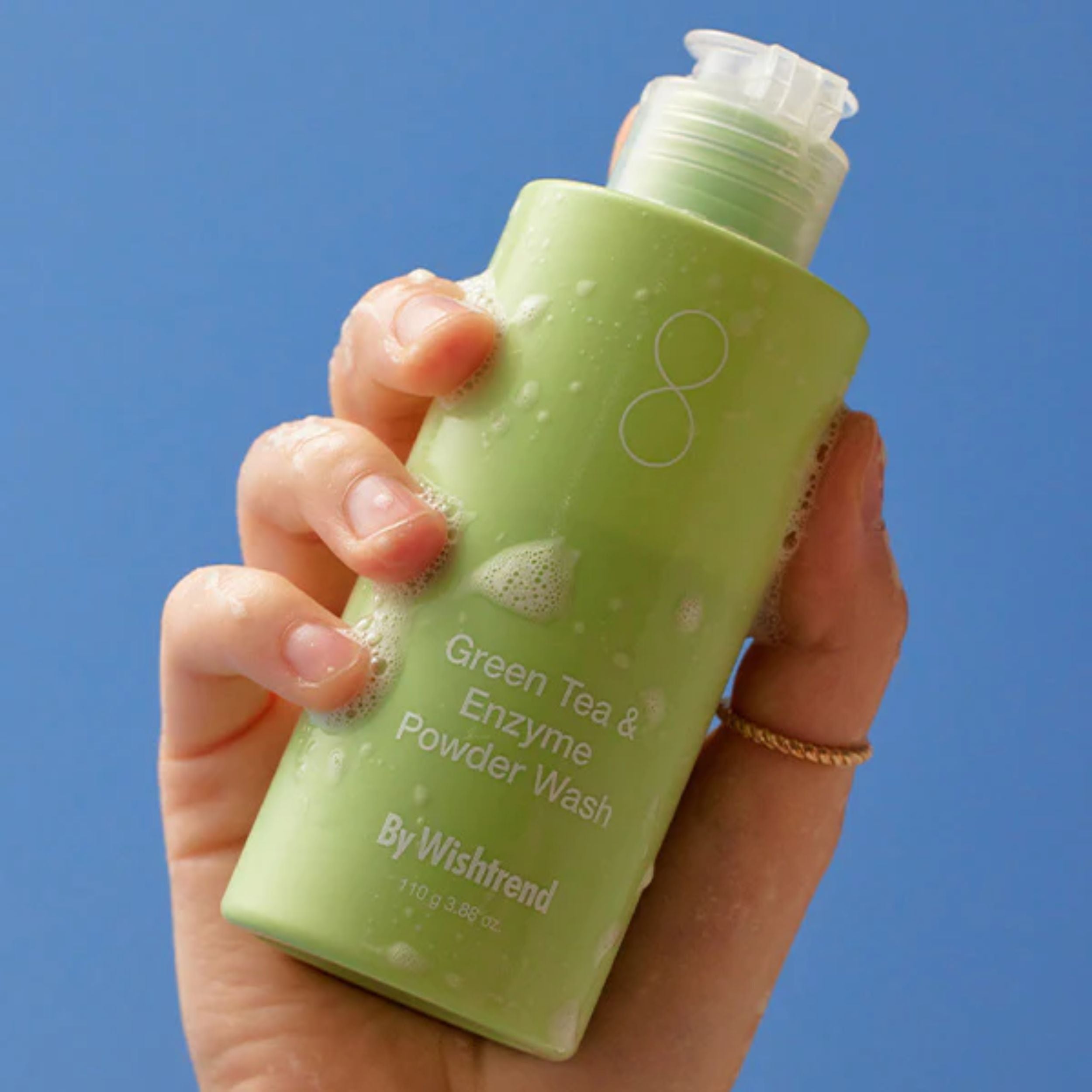 Hand holding a green bottle of 'Green Tea & Enzyme Powder Wash' by Wishtrend against a blue background