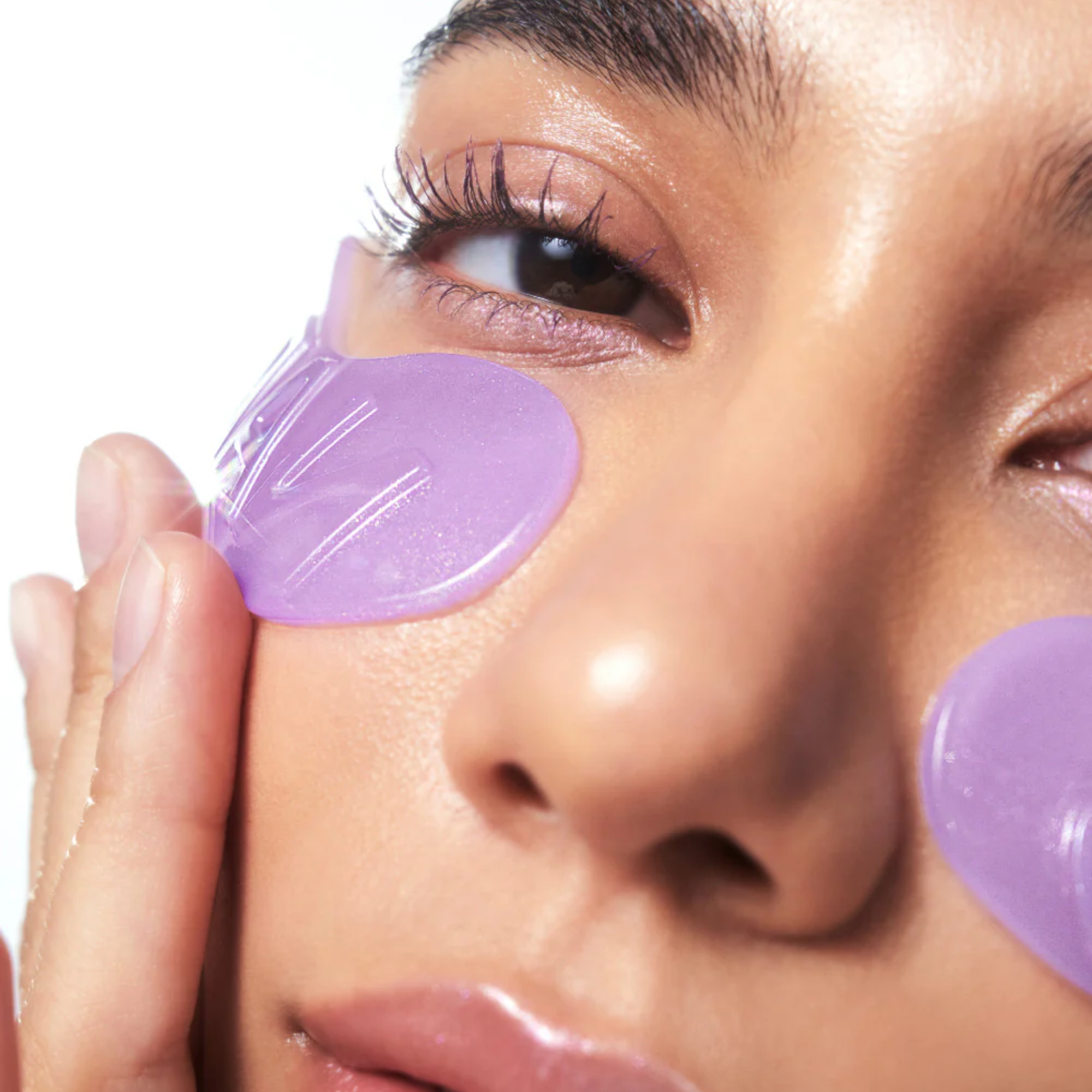 Close-up of a person applying or wearing translucent purple eye patches under their eyes, with one patch being peeled off by a hand against a clean white background.